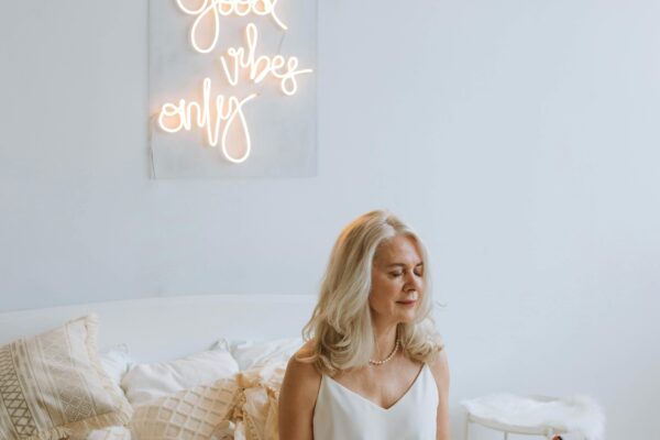 Serene woman meditating on a cozy bed under 'Good Vibes Only' neon sign.